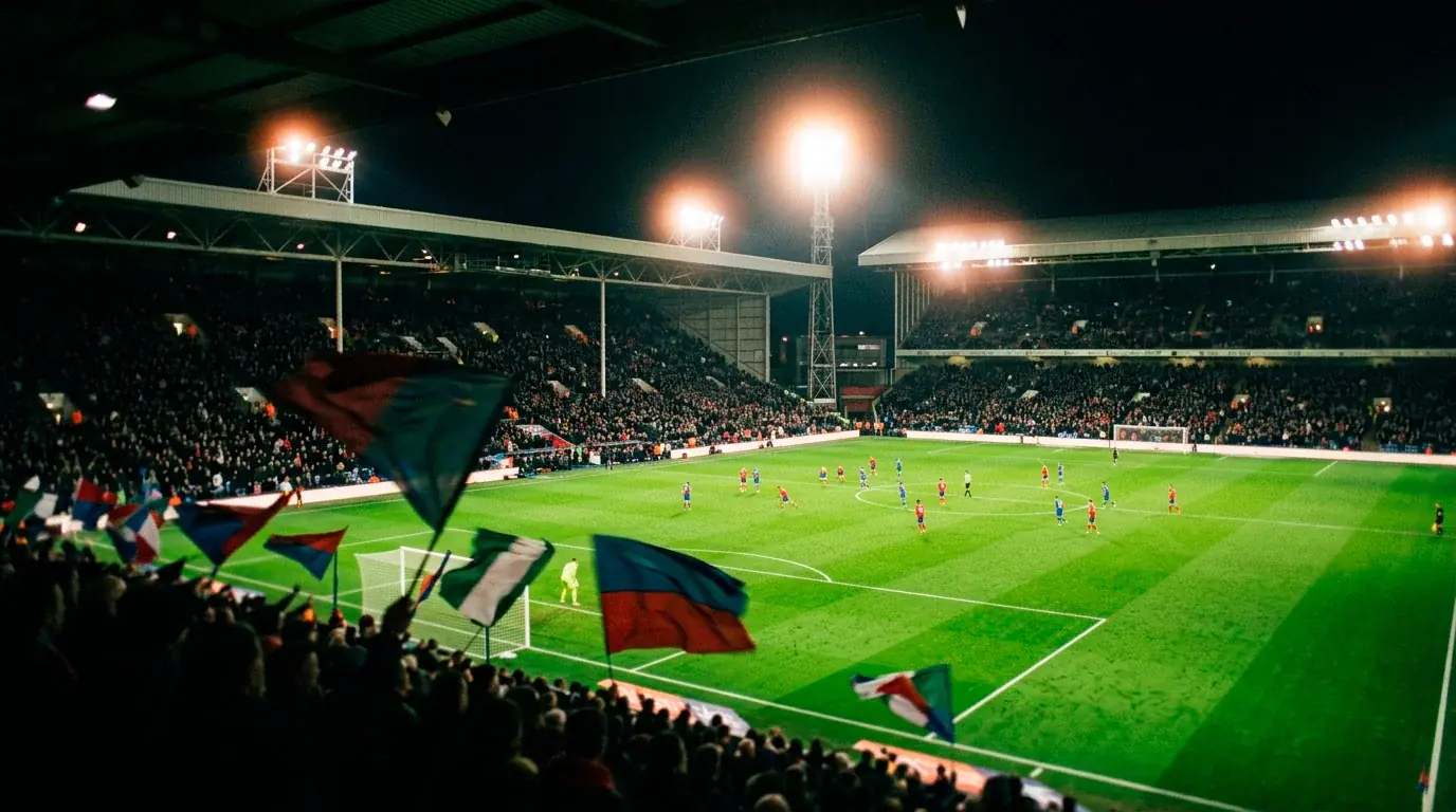 A floodlit football pitch at night with assorted national-team flags blurred in the foreground, viewed from a high stadium angle